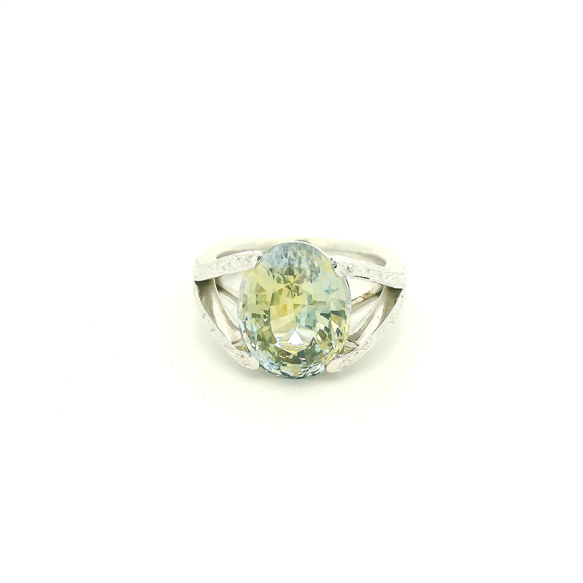 This custom ring features a stunning 9.06 ct oval-cut parti sapphire, showcasing a unique blend of blue, green, and golden tones. Set in 18k white gold, the ring is finished with intricate hand-engraved filigree detailing throughout the split-shank setting, creating a truly one-of-a-kind piece centered around the sapphire’s natural color.