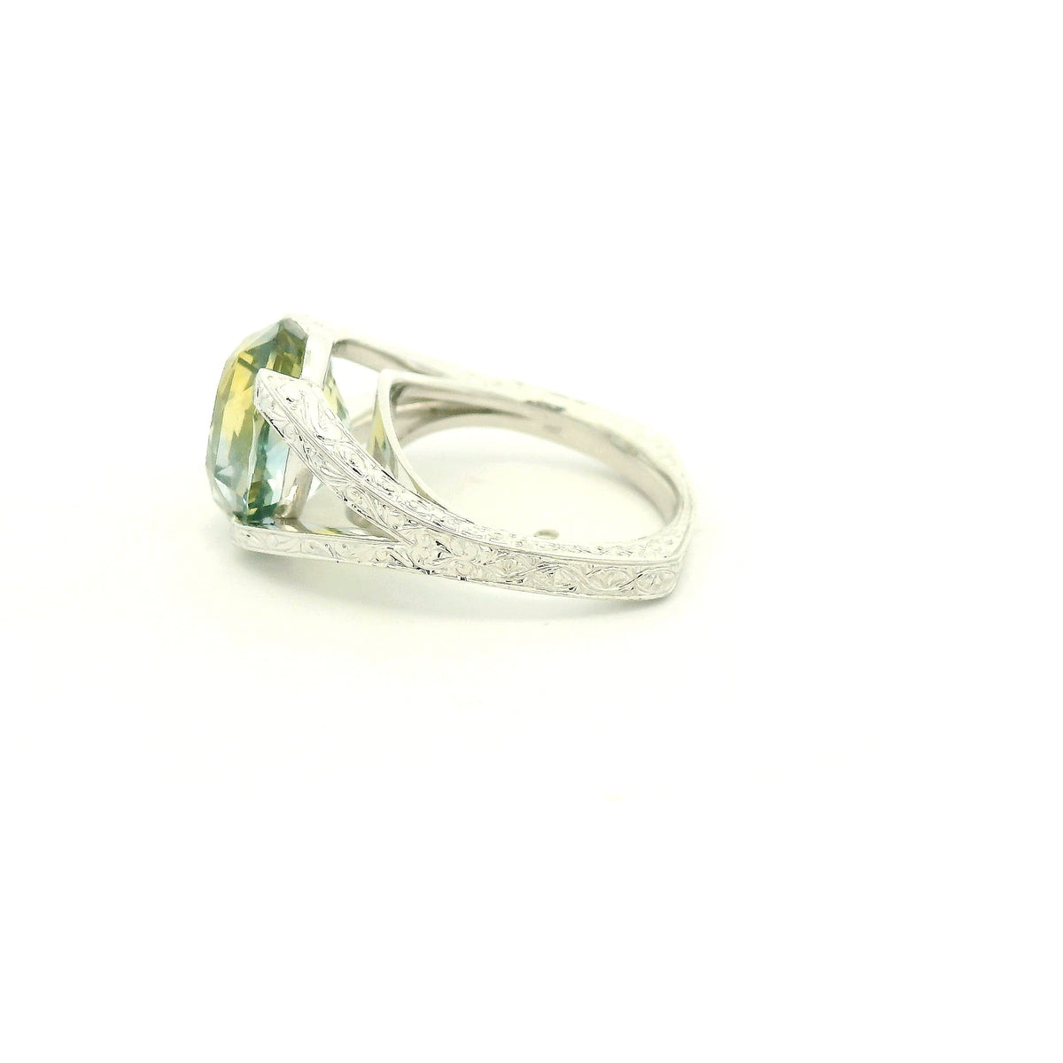 This custom ring features a stunning 9.06 ct oval-cut parti sapphire, showcasing a unique blend of blue, green, and golden tones. Set in 18k white gold, the ring is finished with intricate hand-engraved filigree detailing throughout the split-shank setting, creating a truly one-of-a-kind piece centered around the sapphire’s natural color.