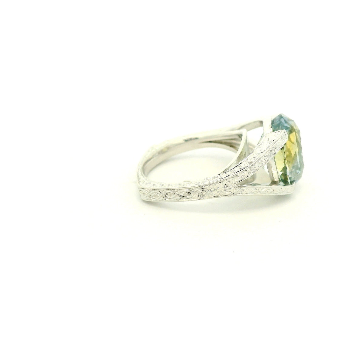 This custom ring features a stunning 9.06 ct oval-cut parti sapphire, showcasing a unique blend of blue, green, and golden tones. Set in 18k white gold, the ring is finished with intricate hand-engraved filigree detailing throughout the split-shank setting, creating a truly one-of-a-kind piece centered around the sapphire’s natural color.