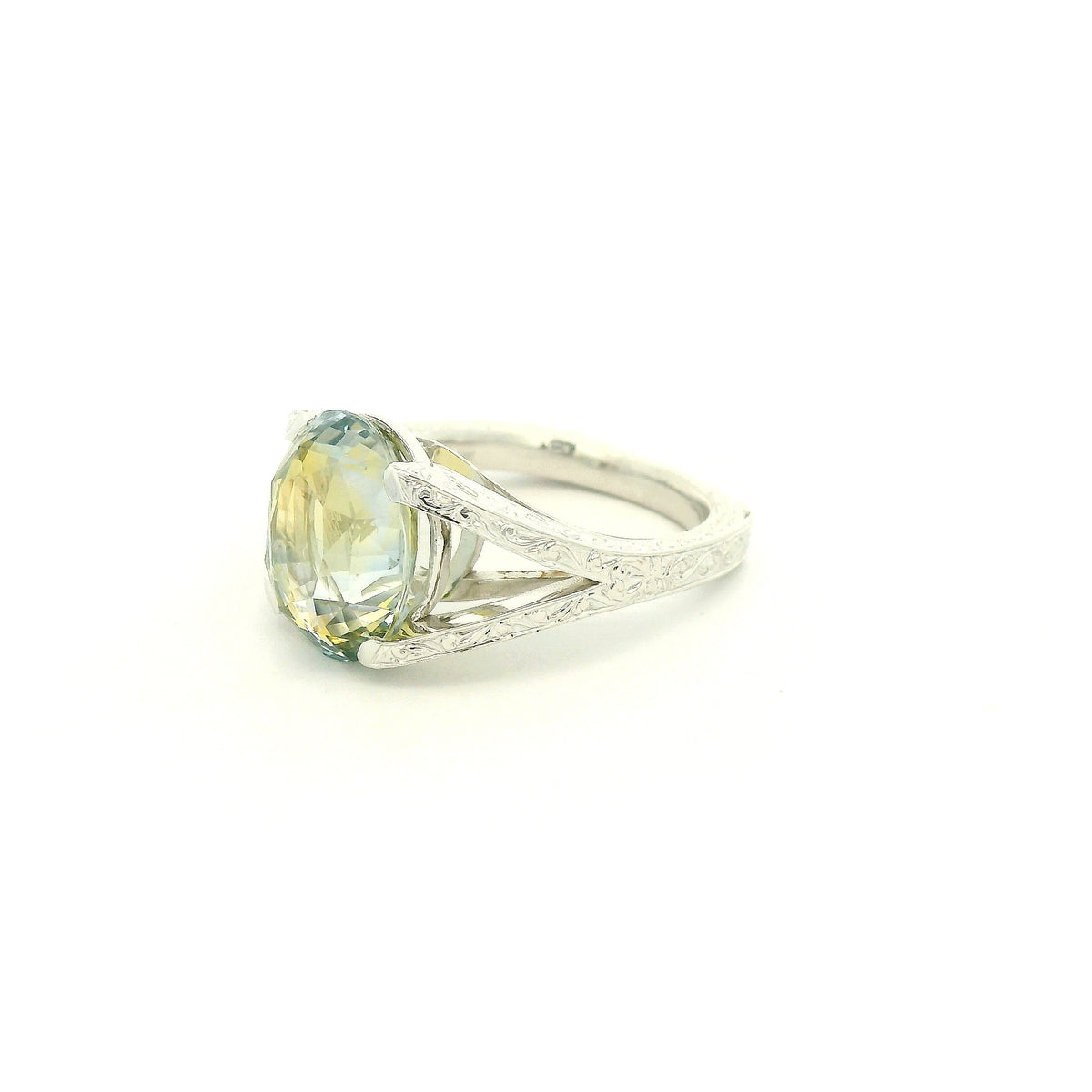 This custom ring features a stunning 9.06 ct oval-cut parti sapphire, showcasing a unique blend of blue, green, and golden tones. Set in 18k white gold, the ring is finished with intricate hand-engraved filigree detailing throughout the split-shank setting, creating a truly one-of-a-kind piece centered around the sapphire’s natural color.