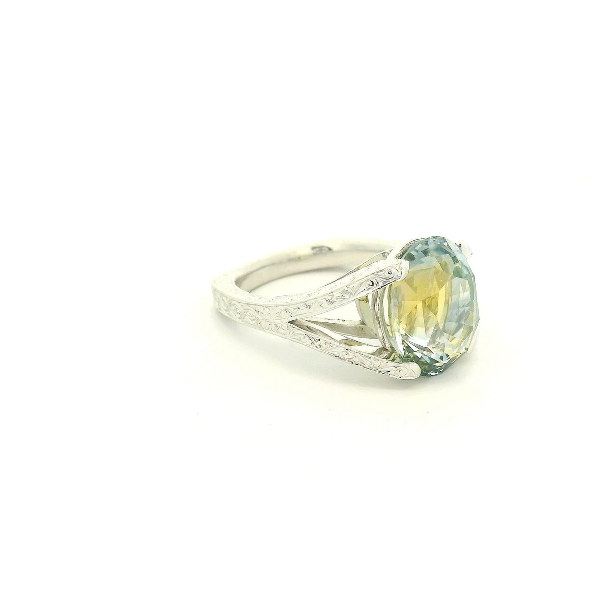 This custom ring features a stunning 9.06 ct oval-cut parti sapphire, showcasing a unique blend of blue, green, and golden tones. Set in 18k white gold, the ring is finished with intricate hand-engraved filigree detailing throughout the split-shank setting, creating a truly one-of-a-kind piece centered around the sapphire’s natural color.