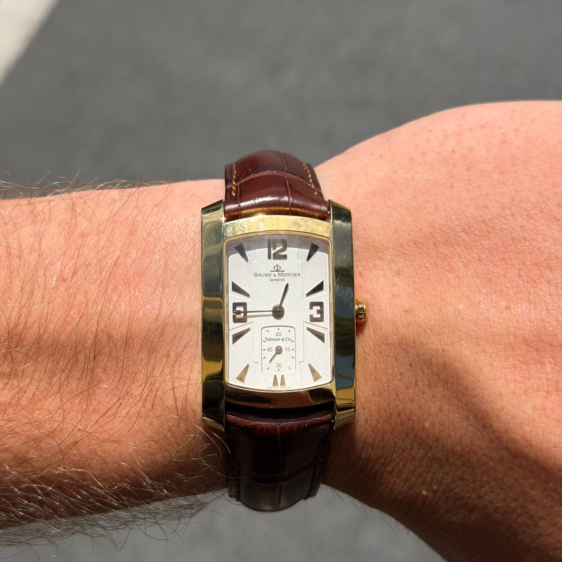 A Vintage Baume & Mercier watch with a 18k gold rectangular case, cream dial with 9, 12 & 3 displayed, the dial is stamped with a tiffany & Co., and dark brown leather strap, shown against a plain white background at Forever Rox Fine Jewelry, Lake Tahoes Best Jewelry Store