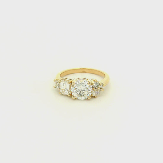 This custom 14k yellow gold engagement ring features a 1.53 ct H-VS2 round brilliant diamond accented by an asymmetrical arrangement of mixed-cut natural diamonds, including a .70 ct F-VVS2 emerald cut, .15 ct G-VS2 pear cut, .17 ct square emerald cut, and two smaller round diamonds. 