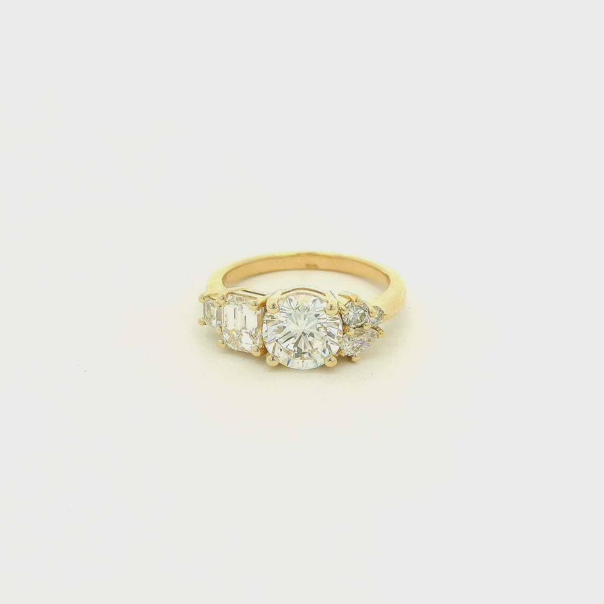 This custom 14k yellow gold engagement ring features a 1.53 ct H-VS2 round brilliant diamond accented by an asymmetrical arrangement of mixed-cut natural diamonds, including a .70 ct F-VVS2 emerald cut, .15 ct G-VS2 pear cut, .17 ct square emerald cut, and two smaller round diamonds. 