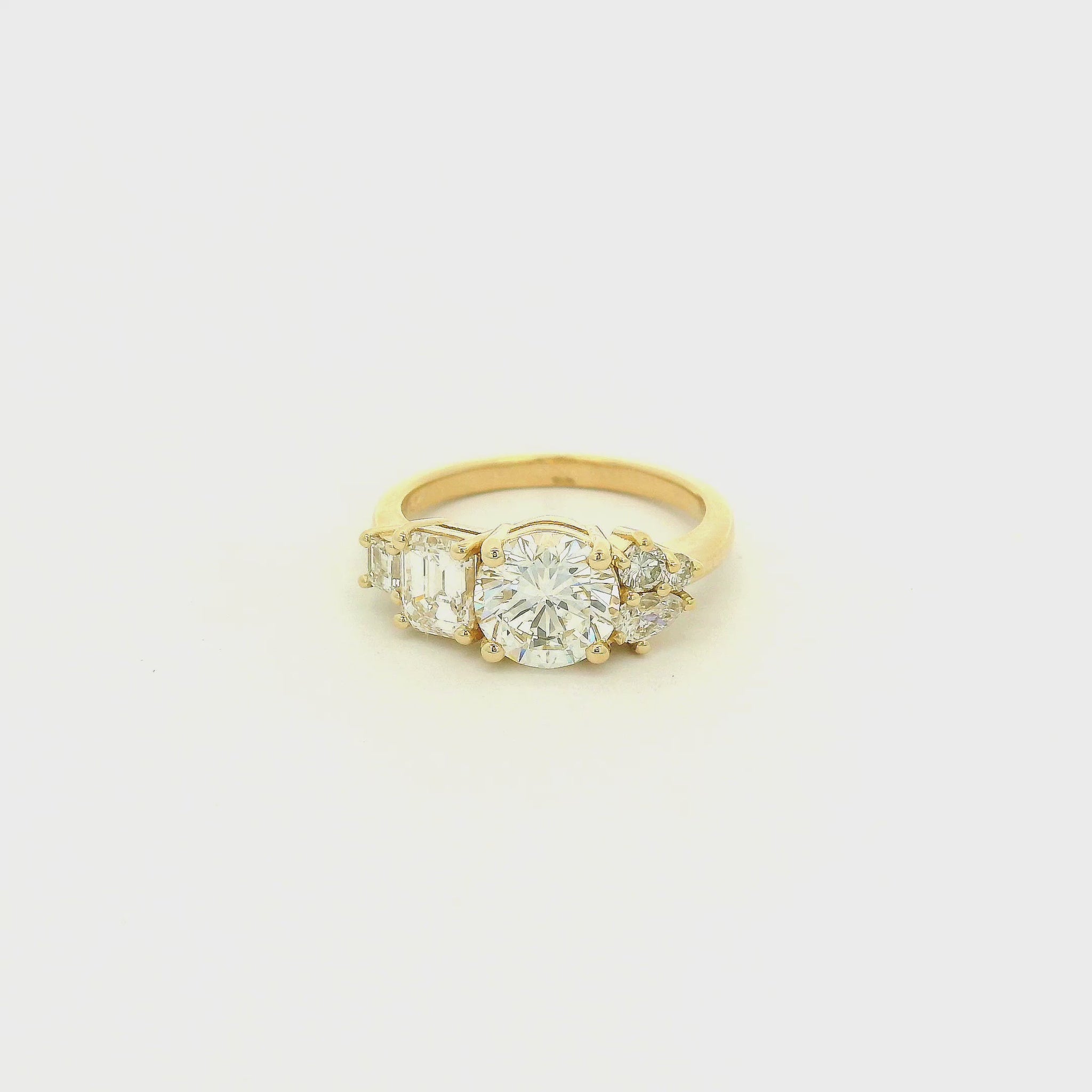 This custom 14k yellow gold engagement ring features a 1.53 ct H-VS2 round brilliant diamond accented by an asymmetrical arrangement of mixed-cut natural diamonds, including a .70 ct F-VVS2 emerald cut, .15 ct G-VS2 pear cut, .17 ct square emerald cut, and two smaller round diamonds. 