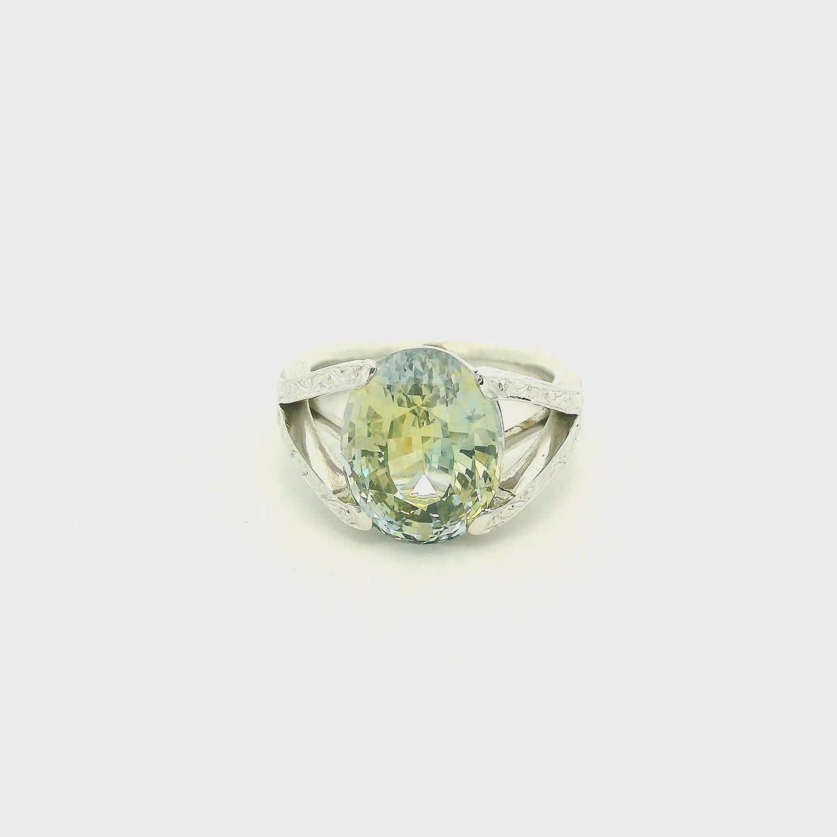 This custom ring features a stunning 9.06 ct oval-cut parti sapphire, showcasing a unique blend of blue, green, and golden tones. Set in 18k white gold, the ring is finished with intricate hand-engraved filigree detailing throughout the split-shank setting, creating a truly one-of-a-kind piece centered around the sapphire’s natural color.