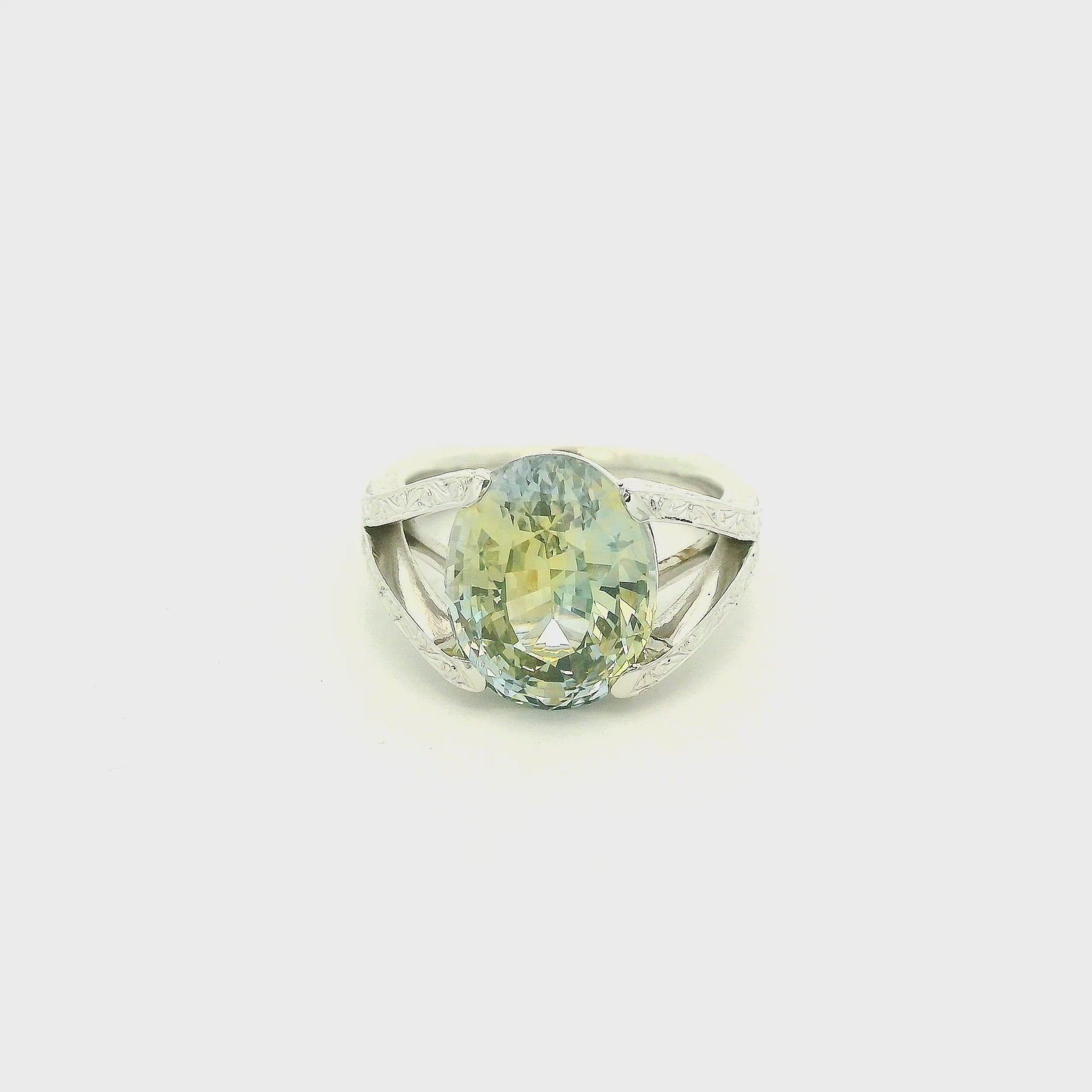 This custom ring features a stunning 9.06 ct oval-cut parti sapphire, showcasing a unique blend of blue, green, and golden tones. Set in 18k white gold, the ring is finished with intricate hand-engraved filigree detailing throughout the split-shank setting, creating a truly one-of-a-kind piece centered around the sapphire’s natural color.